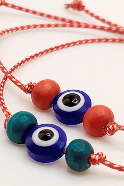 Μαρτάκι με ματόχαντρο (10mm) στο κέντρο, πλαισιωμένο από δύο ξύλινες χάντρες σε κόκκινο ή πετρόλ. Φτιαγμένο από μονό, γυαλιστερό μαρτιάτικο κορδόνι 2mm. Με αυξομειούμενο κλείσιμο ώστε να ταιριάζει σε κάθε καρπό. Σε δύο standard μεγέθη, ανδρικό και γυναικείο, αλλά μπορεί να γίνει και στο ακριβές μέγεθος που θέλετε.