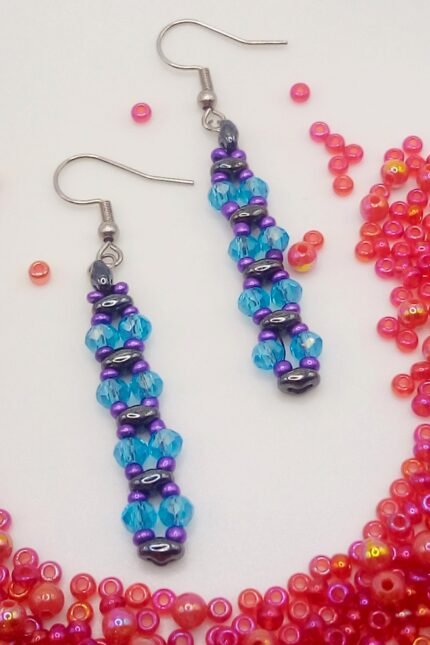 Κρεμαστά σκουλαρίκια μπάρες, φτιαγμένα από χάντρες Superduo στο χρώμα του αιματίτη, συνδυασμένα με γυάλινα κρύσταλλα σε τιρκουάζ. Το σχέδιο συμπληρώνεται από μωβ σατινέ seed beads. Κλείσιμο με γαντζάκι και στοπάκια σιλικόνης. Τα μεταλλικά στοιχεία είναι υποαλλεργικά καθώς είναι φτιαγμένα από χειρουργικό ατσάλι.