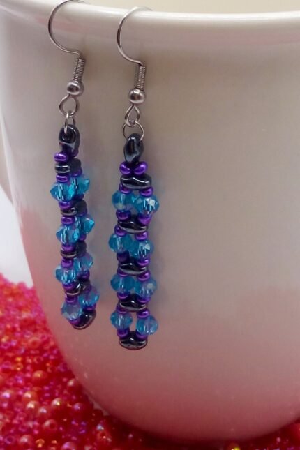 Κρεμαστά σκουλαρίκια μπάρες, φτιαγμένα από χάντρες Superduo στο χρώμα του αιματίτη, συνδυασμένα με γυάλινα κρύσταλλα σε τιρκουάζ. Το σχέδιο συμπληρώνεται από μωβ σατινέ seed beads. Κλείσιμο με γαντζάκι και στοπάκια σιλικόνης. Τα μεταλλικά στοιχεία είναι υποαλλεργικά καθώς είναι φτιαγμένα από χειρουργικό ατσάλι.