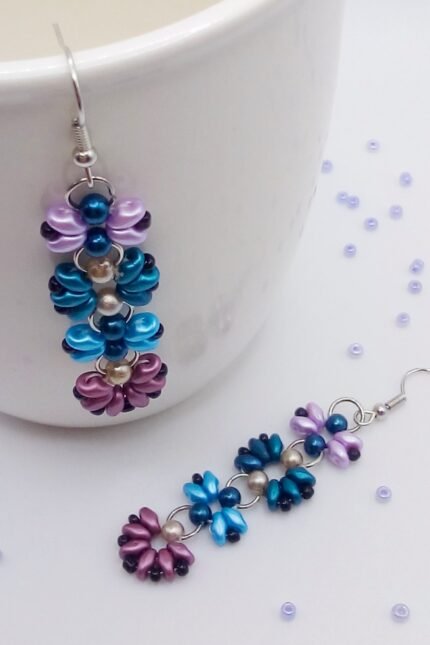 Μοντέρνα, κρεμαστά σκουλαρίκια μπάρες, φτιαγμένα από χάντρες Superduo σε σχέδιο λουλούδια και πεταλούδες. Σε έναν ενδιαφέροντα συνδυασμό πετρόλ, γαλάζιο, μωβ και ροζ με μαύρες λεπτομέρειες. 