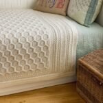 Πλεκτή κουβέρτα με ανάγλυφο μοτίβο κυψέλης σε ανοιχτό μπεζ χρώμα, στρωμένη πάνω σε κρεβάτι με διακοσμητικά μαξιλάρια.