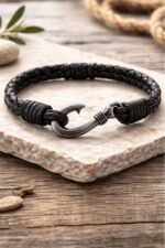 Men’s black braided leather bracelet featuring a single zamak hook charm in gunmetal finish, displayed on stone and wood background.